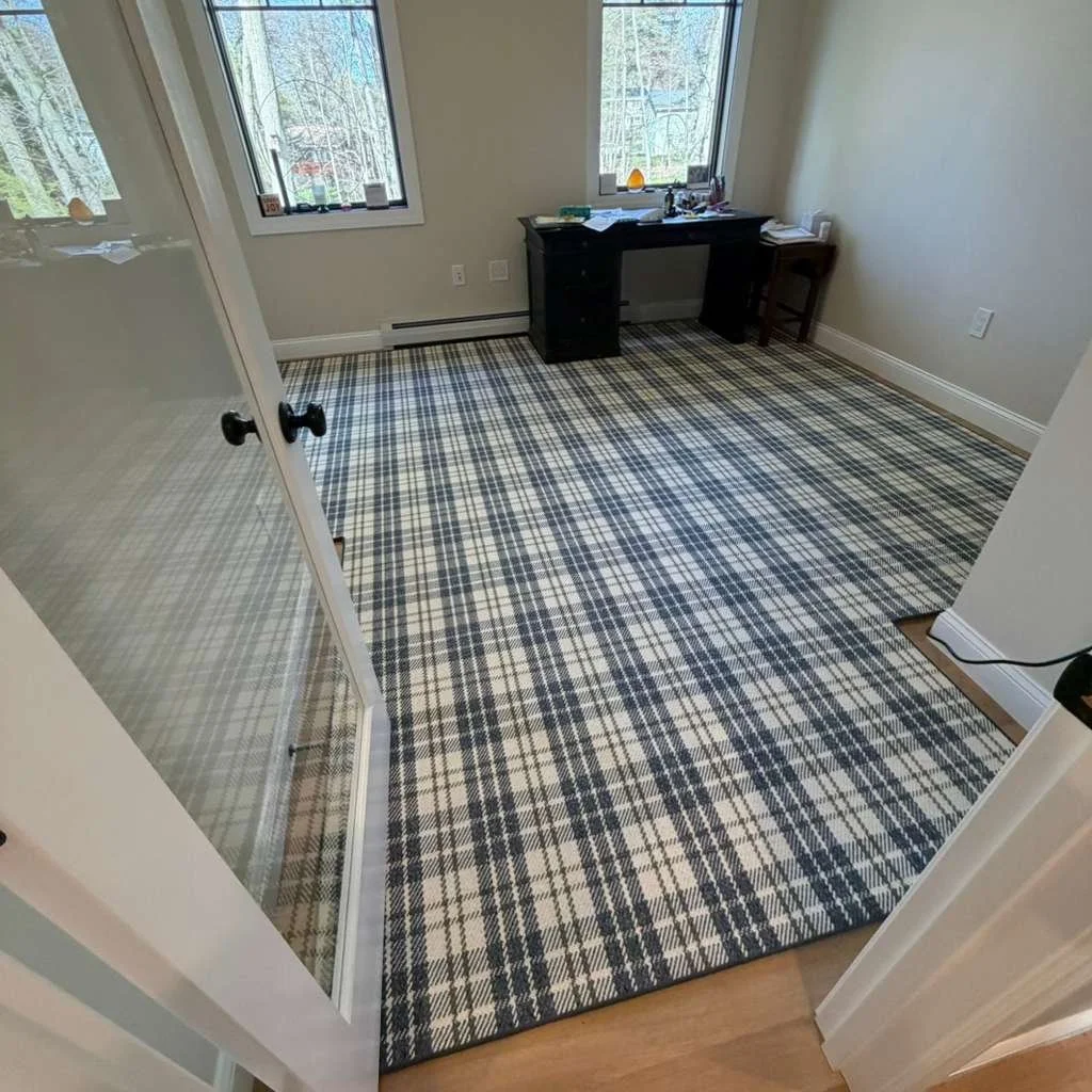 Empty room with plaid patterned carpet, two windows, a black desk with papers and objects, and a small stool beside it.