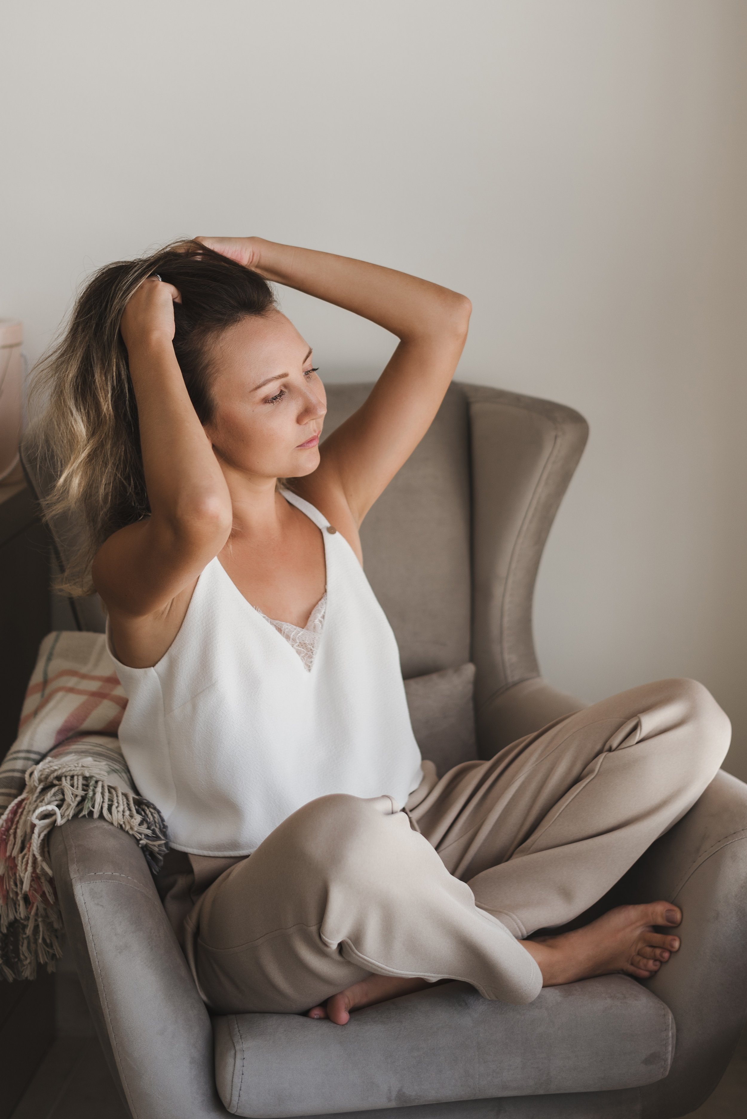 A woman sitting with legs crossed on an armchair, stretching her arms behind her head, wearing a white sleeveless top and beige pants, with a relaxed expression.