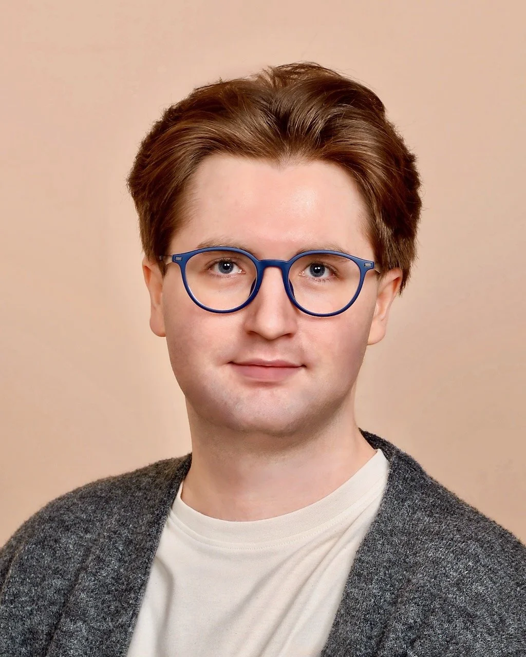 A young man with brown hair and blue glasses, wearing a white shirt and dark gray sweater, against a neutral beige background.