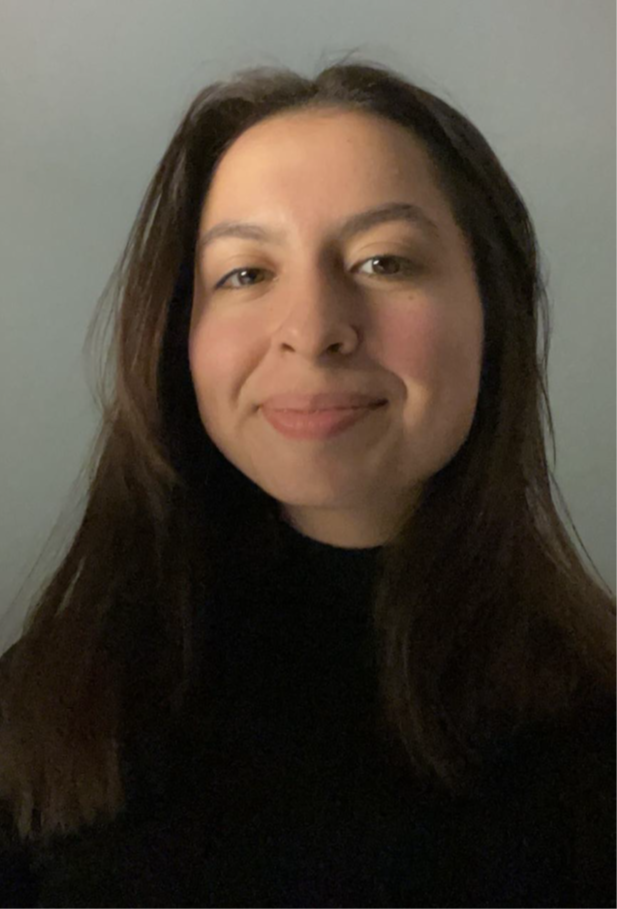 A woman with long, dark hair smiling and wearing a black top against a plain wall background.