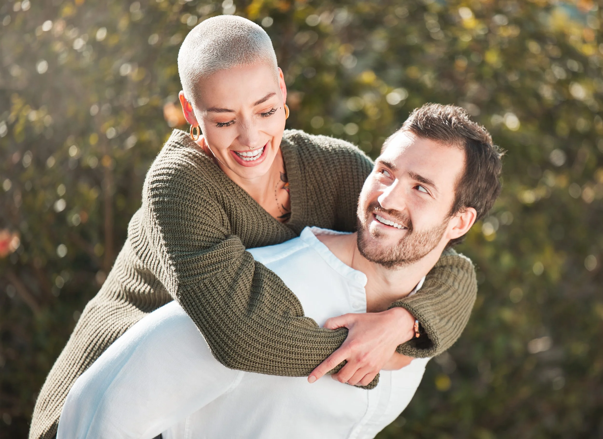 A woman with a buzz cut haircut and earrings playfully piggybacking a smiling man outdoors, with sunlight and blurred trees in the background.