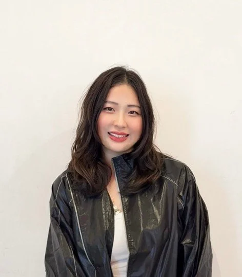 Young woman with wavy dark hair smiling, wearing a black leather jacket over a white top, standing against a plain white wall.