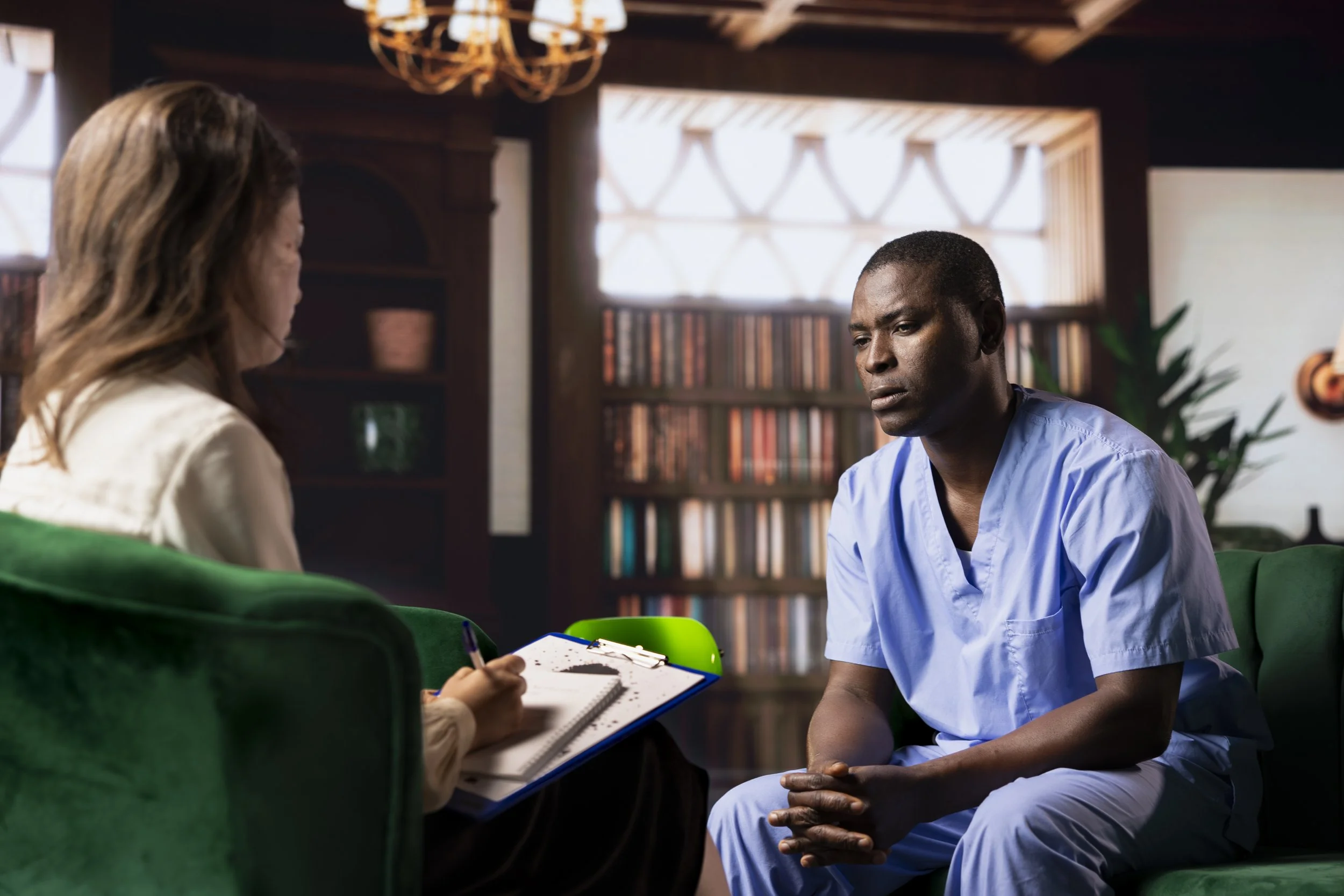 A man in light blue medical scrubs sitting on a green couch during a therapy session with a therapist taking notes.