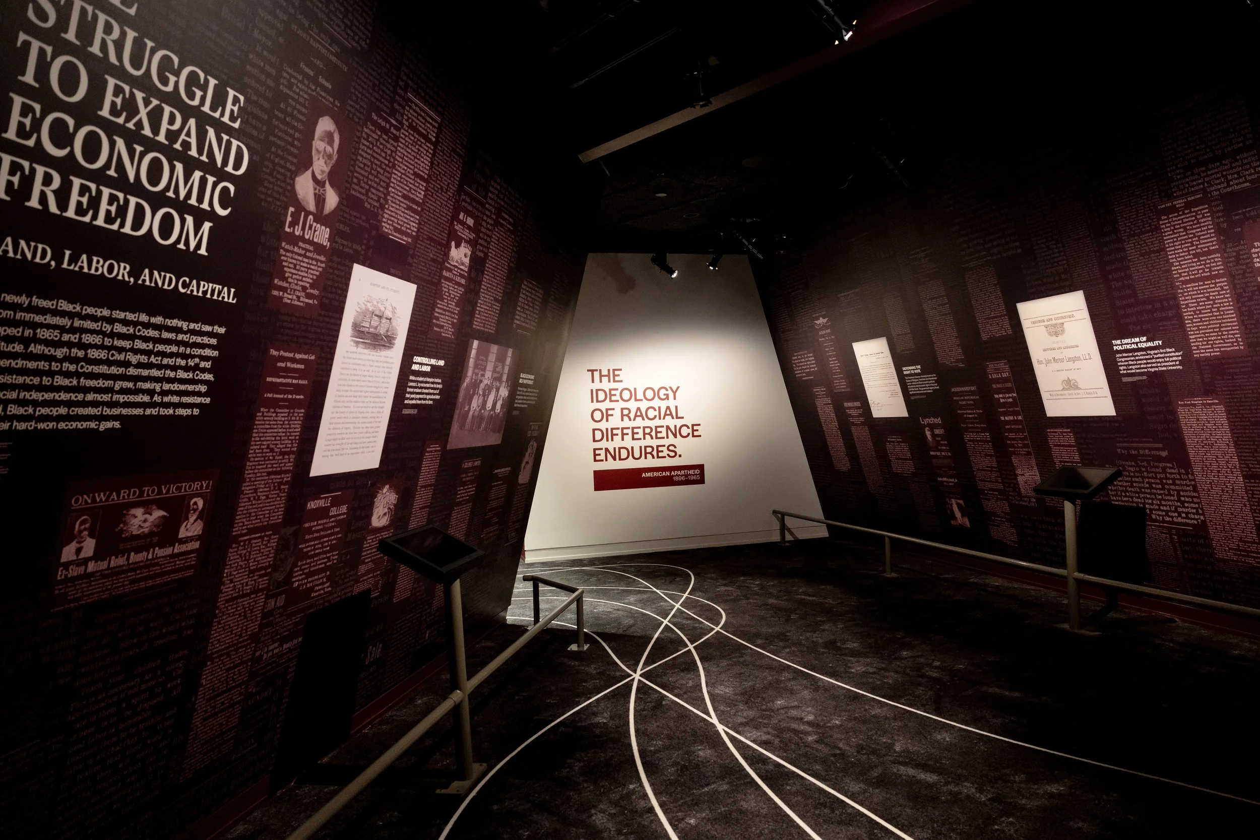 An exhibition room with maroon walls covered in historical texts, images, and articles, centered around themes of racial inequality and history, including a white wall with the phrase 'The Ideology of Racial Difference Endures' in red text.
