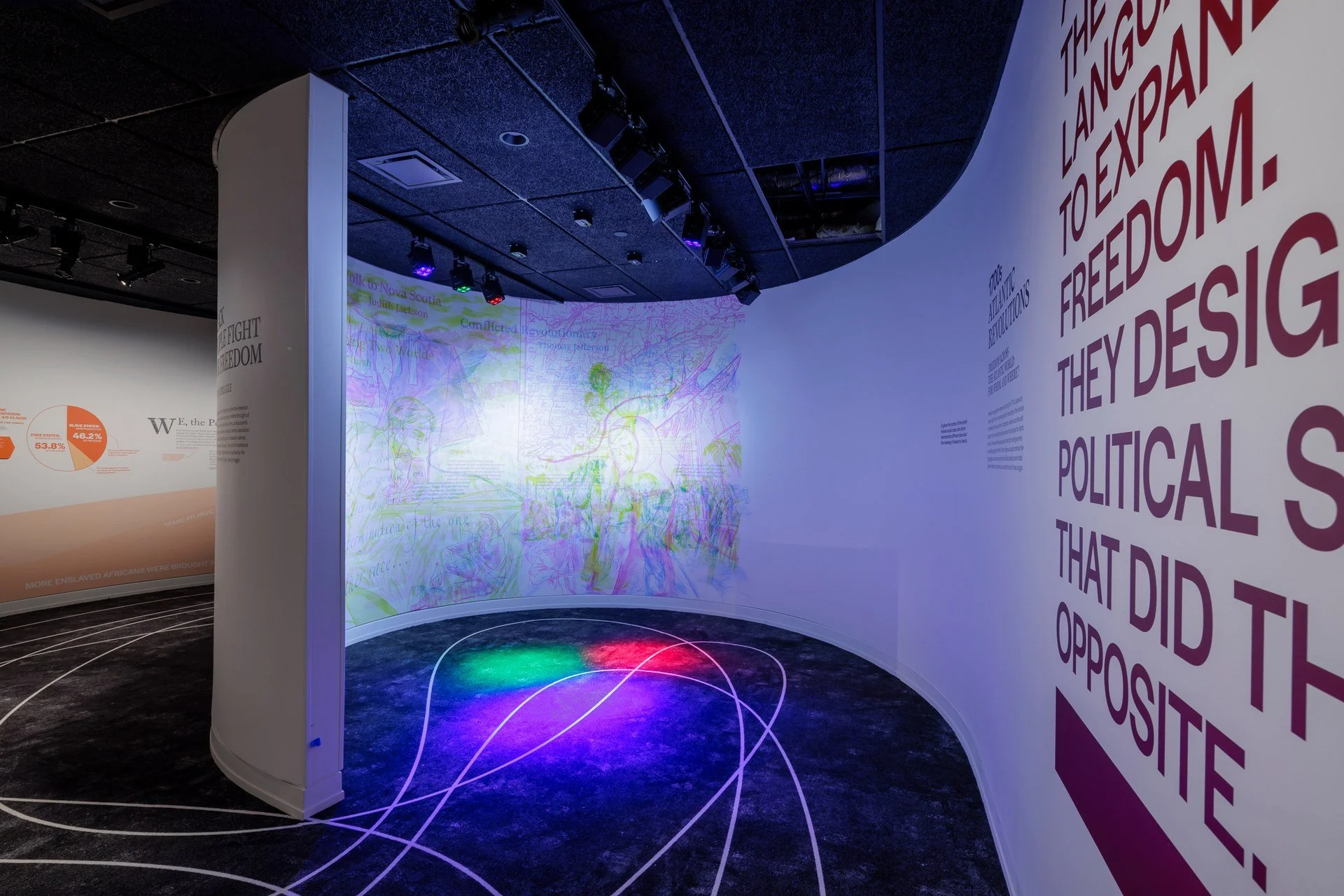 An art exhibit with curved white walls, dark ceiling with multiple lights and speakers, a wall with colorful drawings, and text on another wall that discusses freedom and political themes. There are glowing color lines on the floor.