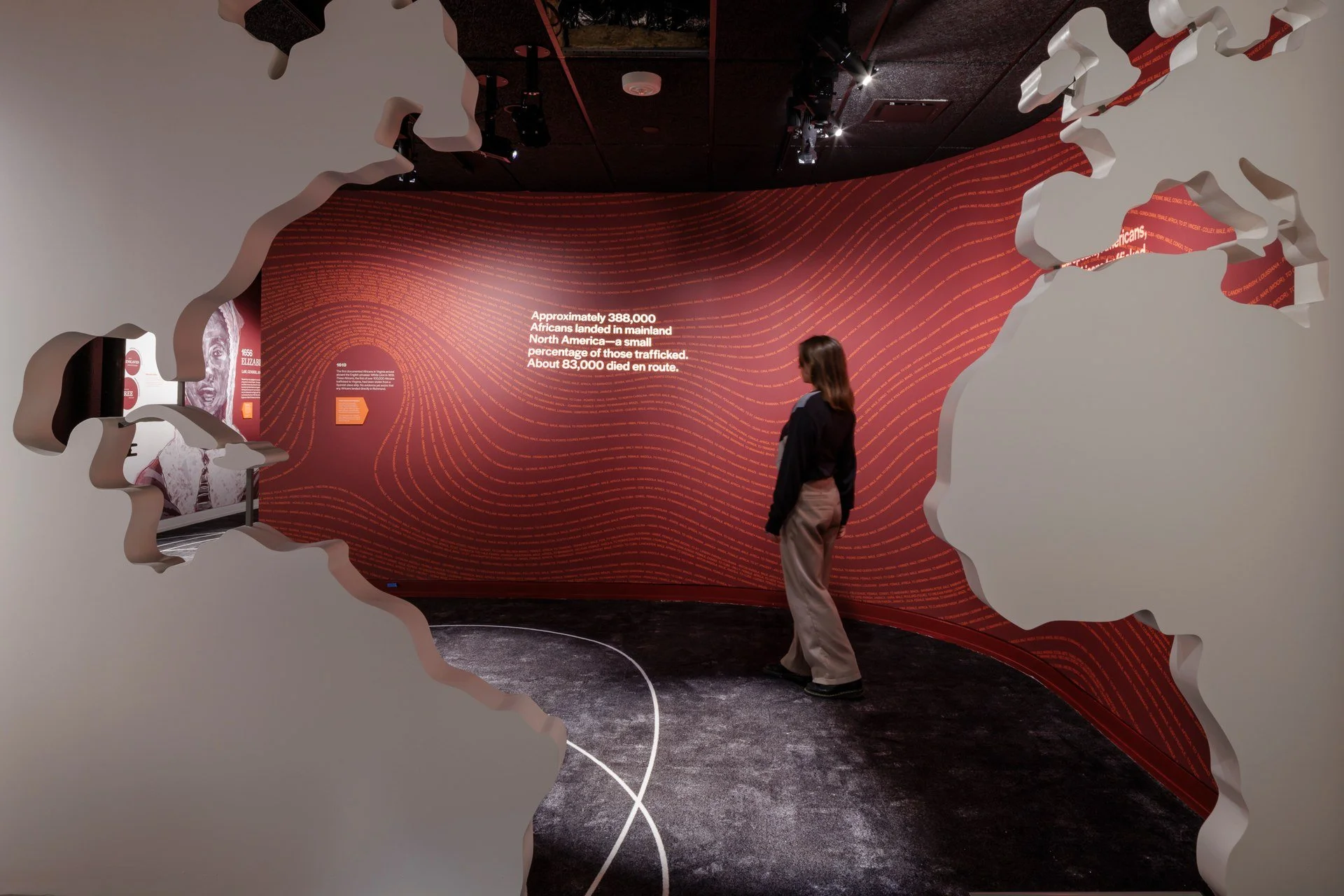 A woman stands inside a circular exhibit with jagged white wall edges, viewing a red wall with text about African migration statistics during the transatlantic slave trade.