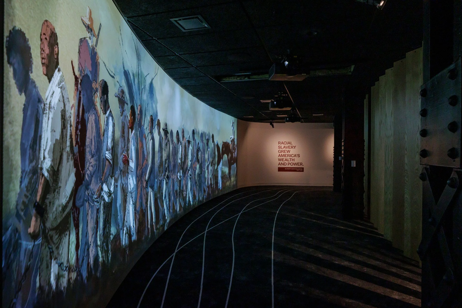 A curved wall with a large mural depicting a diverse group of people in various styles of clothing. The room has dark flooring with white curved lines and a small poster at the end of the wall reads, 'Racial slavery grew America's wealth and power.'