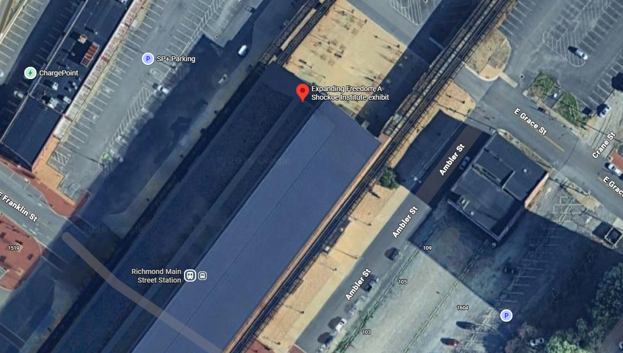 Satellite view of a city block showing a street, parking lots, a train station, and an exhibit titled 'Expanding Freedom, A Shockoe Institute exhibit' near rail tracks.