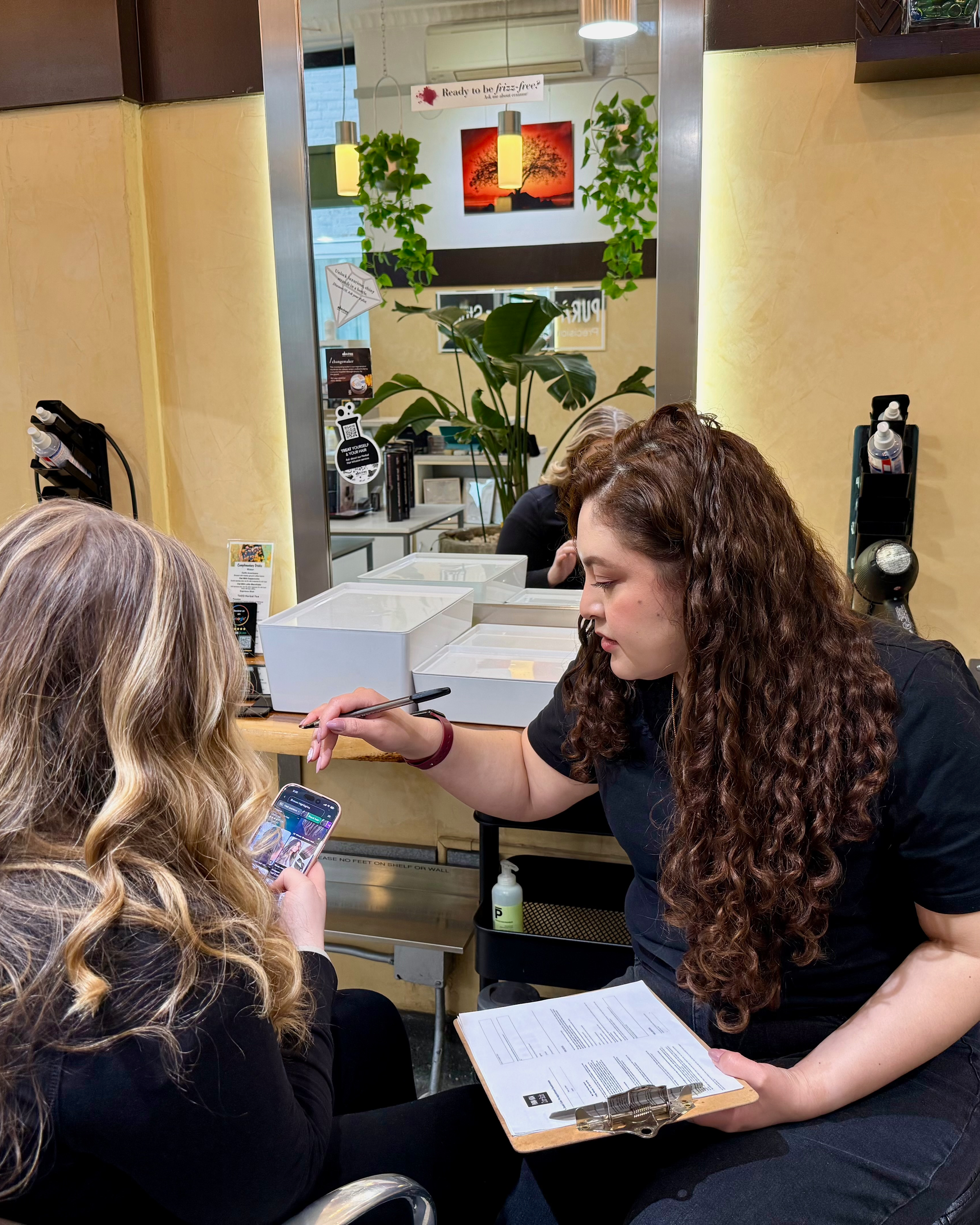 A woman with curly brown hair and a black shirt is holding a clipboard and using a pen on a woman's shoulder. The woman with blonde hair with highlights is holding a smartphone, showing her the screen. They are seated in a salon or spa with a mirror and decorative plants in the background.