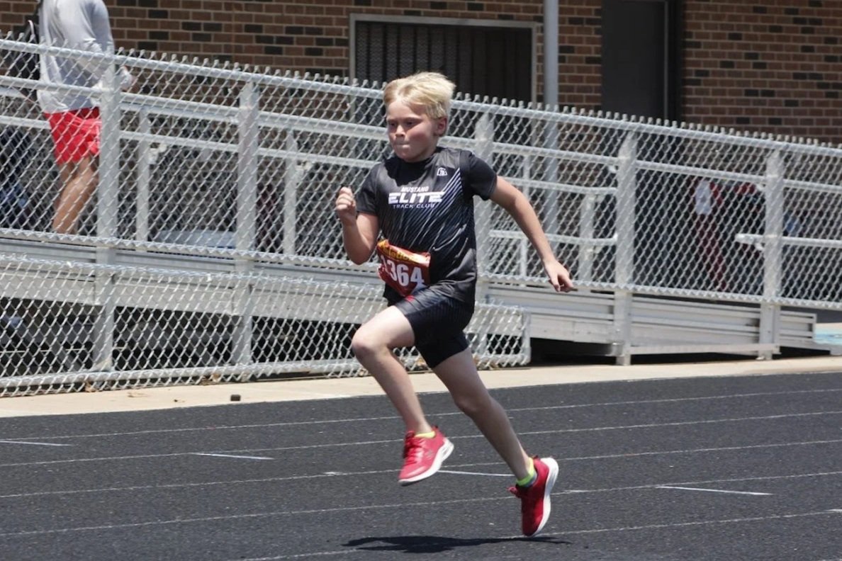 Young boy running on a track during a race, wearing a black athletic shirt, black shorts, pink shoes, and a race bib numbered 364. There are bleachers and a brick building in the background.