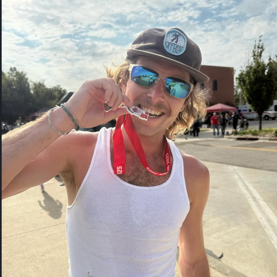 A young man with long hair, wearing sunglasses, a baseball cap, and a white tank top, holding up a finisher’s medal at an outdoor event, with a partly cloudy sky and a few people in the background.