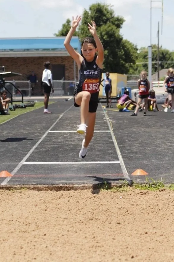 Young female athlete mid-jump during a long jump competition on a track and field day.