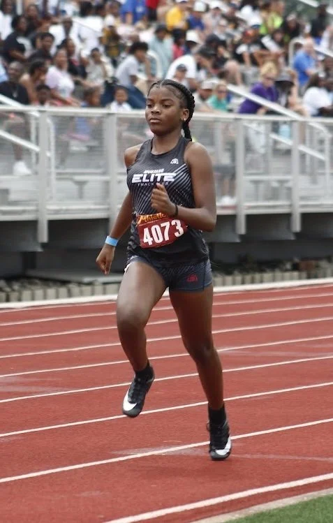 A young female athlete running on a track at a race event, wearing a black sports outfit with a race bib number 4073, with a crowd of spectators in the background.