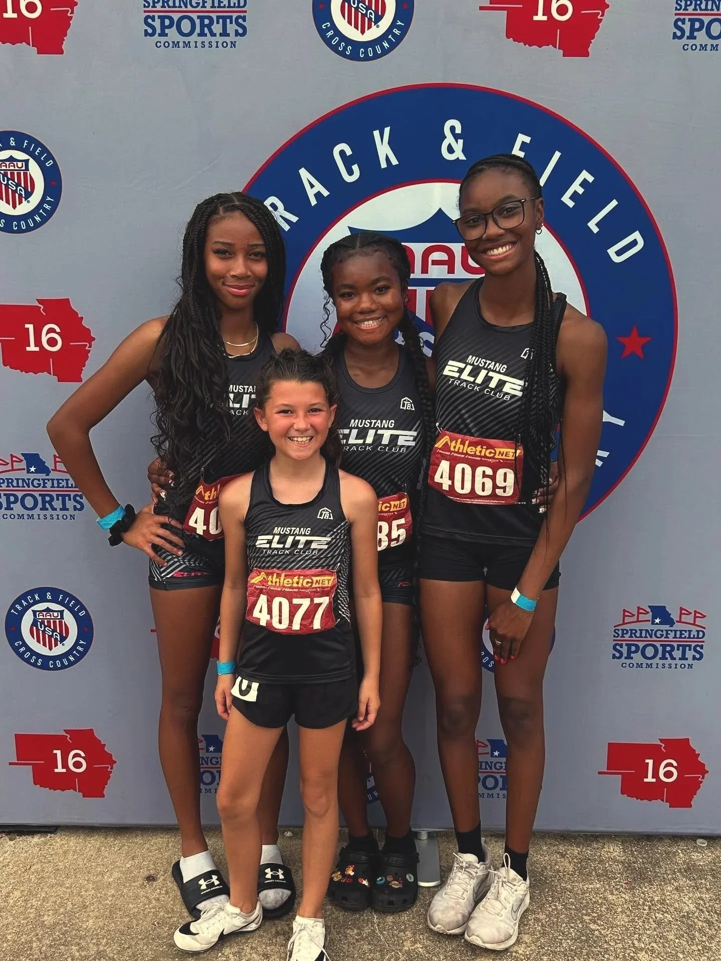 A group of five young female runners in black athletic uniforms with 'Mustang Elite Track Club' printed on them, posing together and smiling in front of a large backdrop with the AAU track and field logo