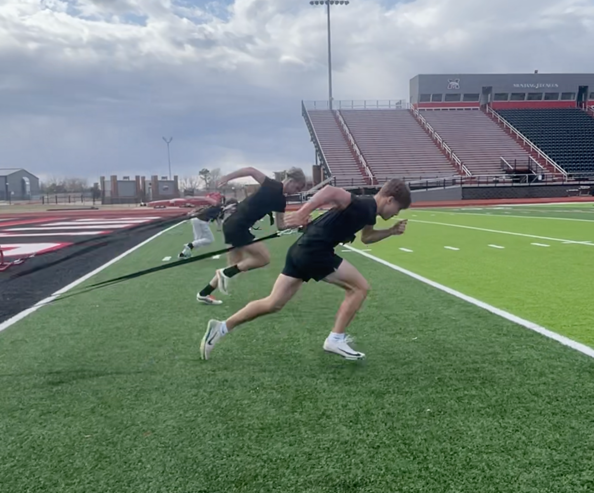 Three athletes sprint on a track at a stadium, with overcast skies in the background.