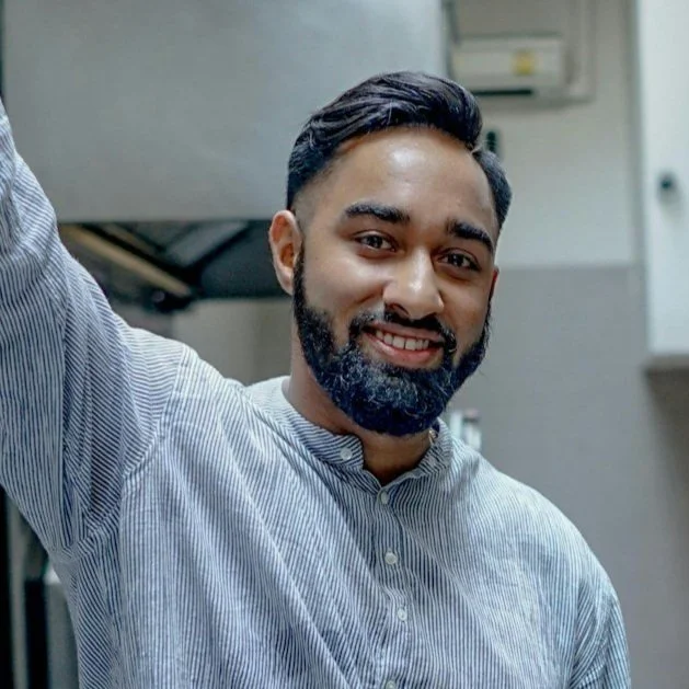 A young man with a beard and short hair smiling in a casual setting, wearing a light blue and white striped shirt.