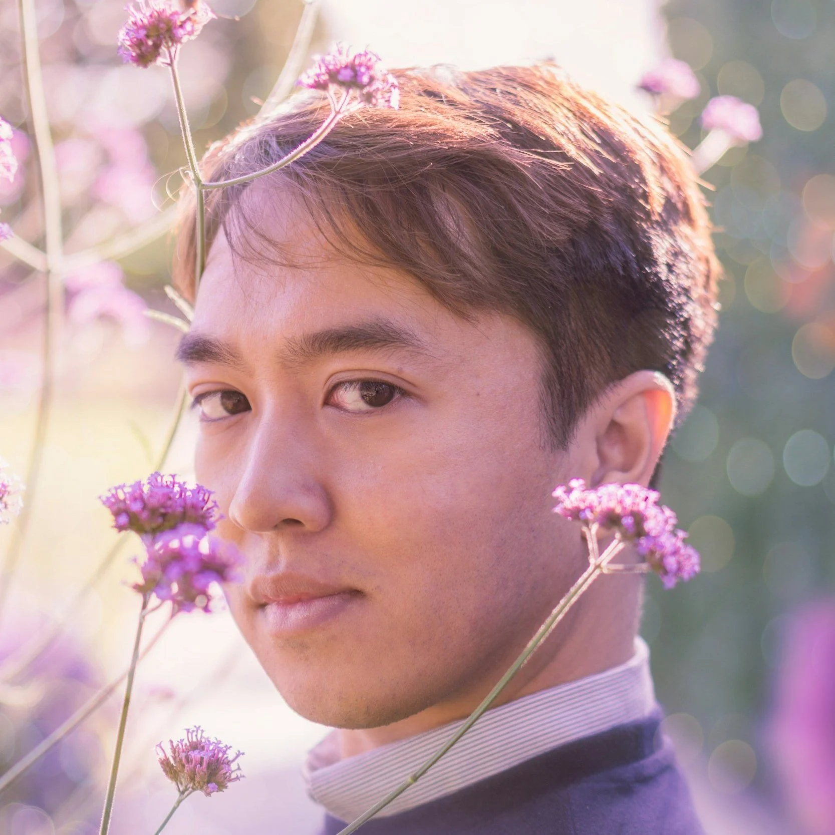 A young man with brown hair and light skin gazes into the camera, surrounded by pink flowers and soft sunlight creating a dreamy bokeh background.