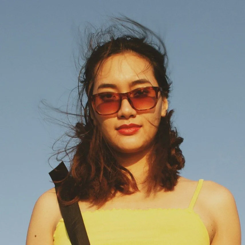 Young woman with wavy brown hair wearing sunglasses and a yellow spaghetti strap top, outdoors with a clear blue sky.