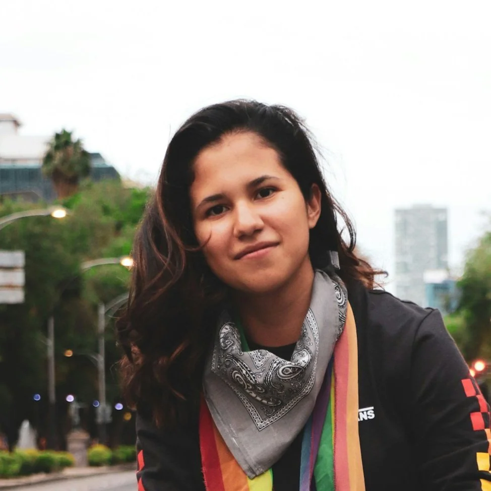 Young woman with dark wavy hair smiling outdoors, wearing a gray bandana and a rainbow-striped scarf, with trees and city buildings in the background.