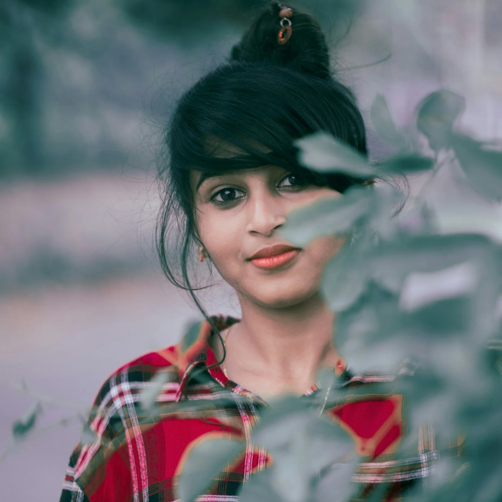 A woman with dark hair styled in a bun, partially obscured by leaves, wearing a red and black plaid shirt and red lipstick, outdoors with blurred trees in the background.