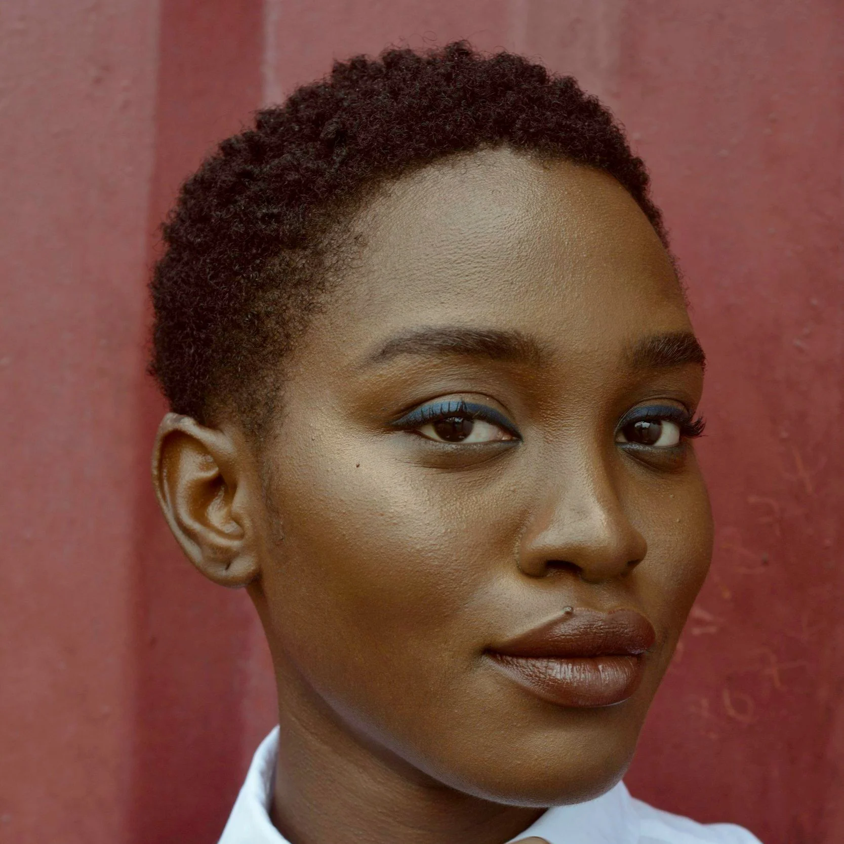 Close-up of a woman with short curly hair, wearing blue eyeliner, dark lipstick, and a white shirt against a reddish background.