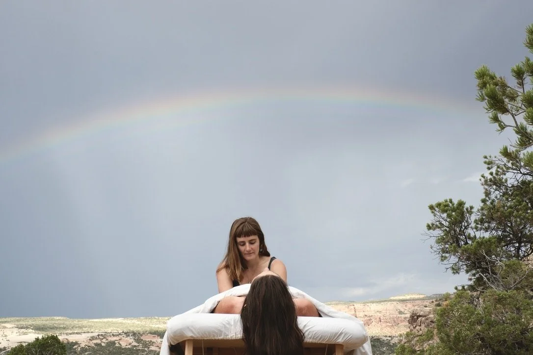 A young woman giving a massage outdoors under a rainbow in a rural landscape with trees and a cloudy sky.