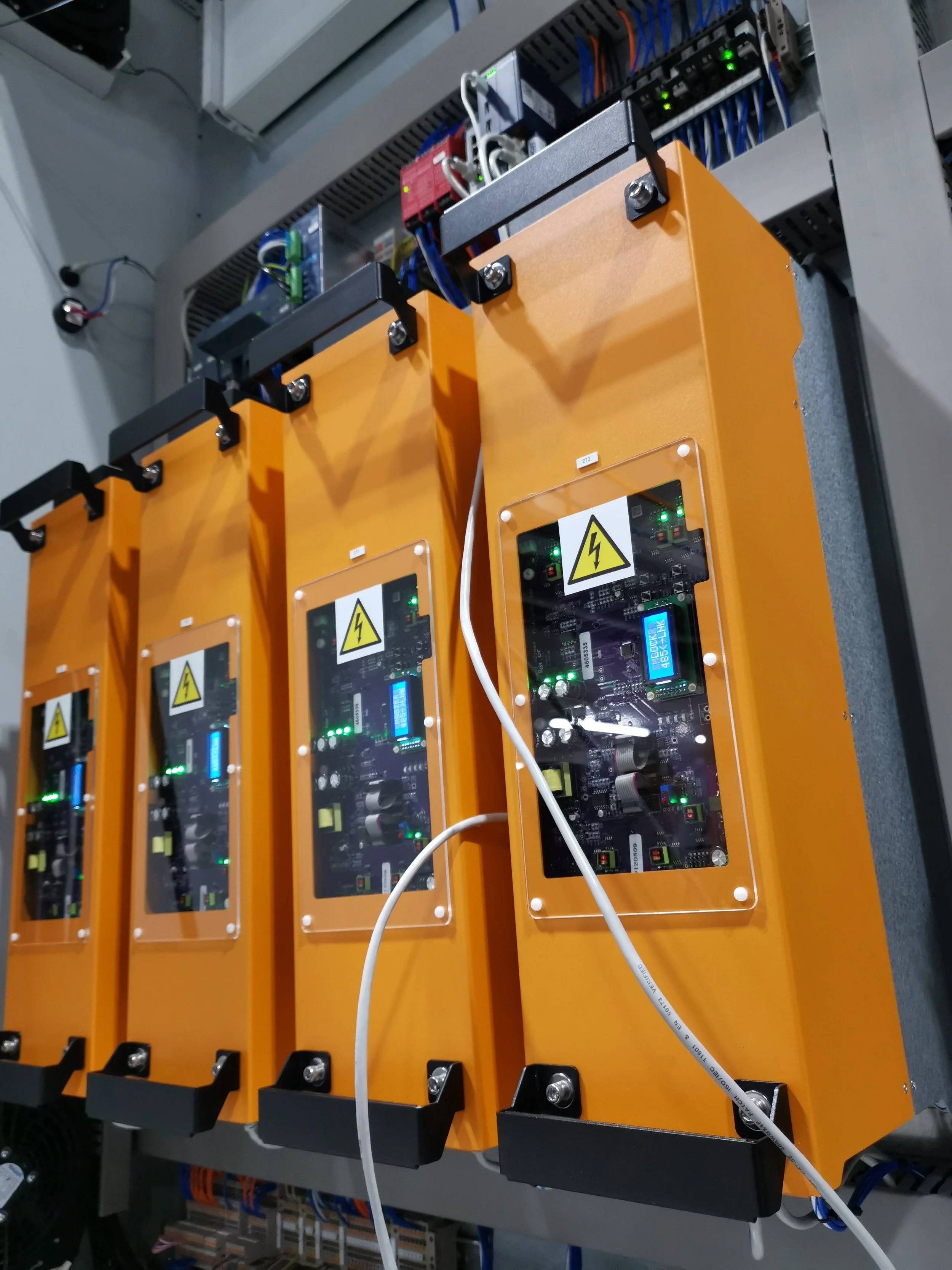 Close-up of three industrial electronic control units enclosed in orange metal cases with transparent covers, mounted on a gray panel, with wires and circuit boards visible inside.