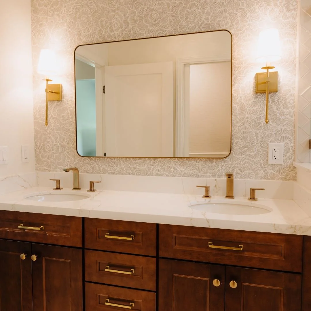 Sophisticated symmetry meets serene design. This master bath continues the story of luxury craftsmanship &mdash; custom wood cabinetry, soft gold finishes, and textured floral wallpaper come together for a timeless yet modern retreat. Every detail, f