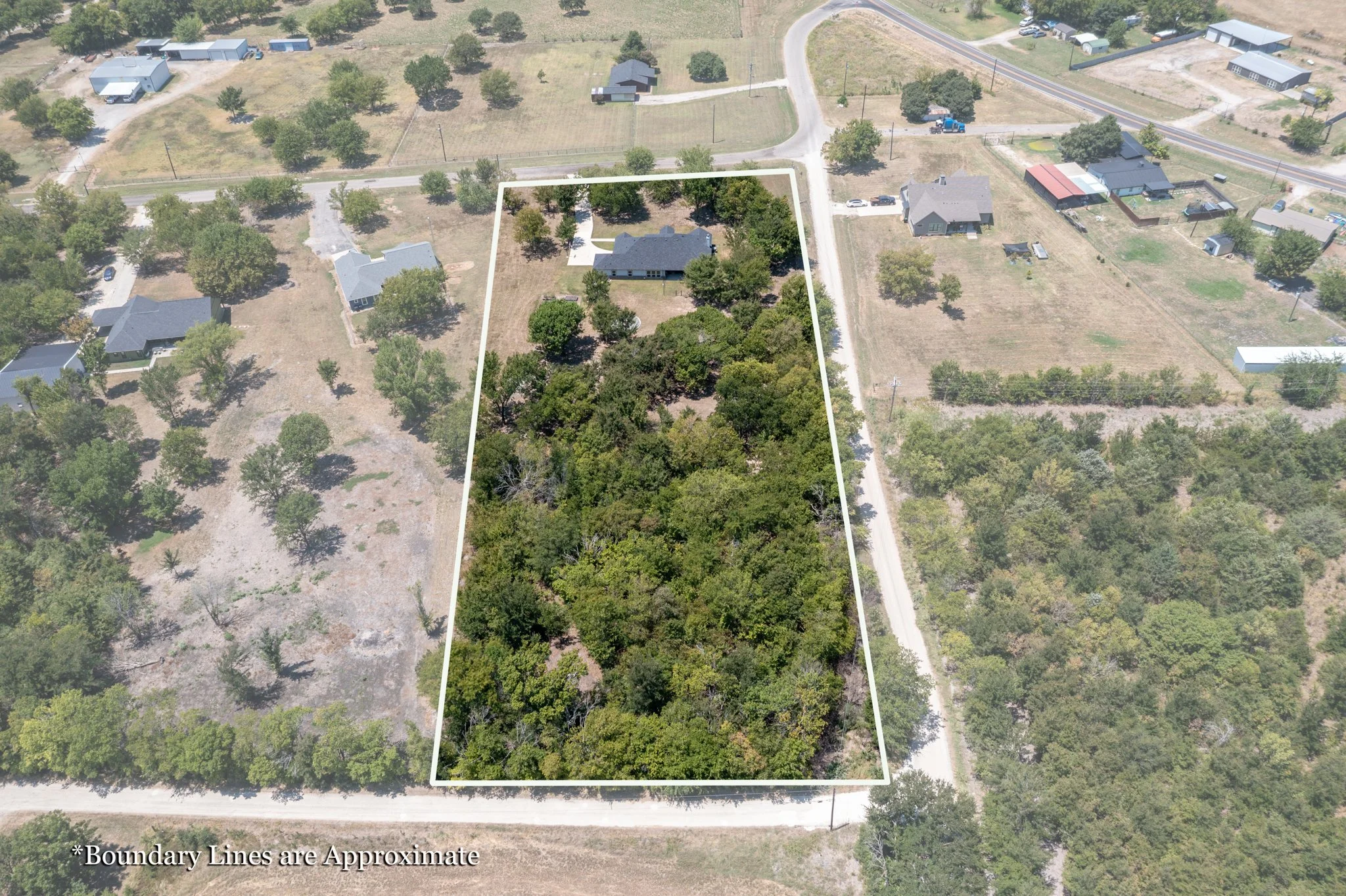 Aerial view of a suburban neighborhood featuring a large yard with numerous trees, outlined by boundary lines, surrounded by several houses, roads, and scattered smaller structures.