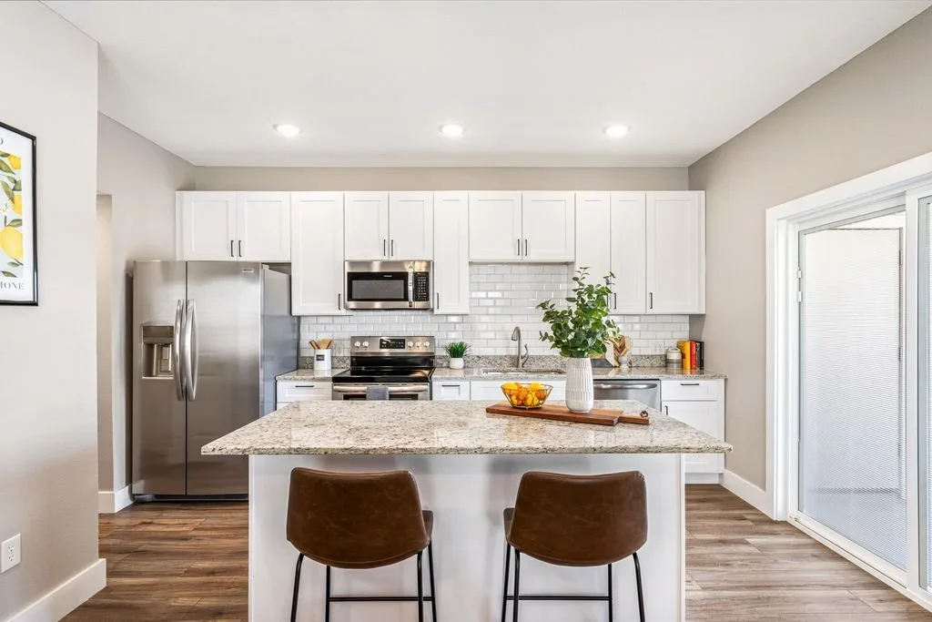 Modern kitchen with white cabinets, stainless steel refrigerator, microwave, stove, granite countertops, and a small island with two brown chairs. There's a sliding glass door on the right and decorative items on the counter.