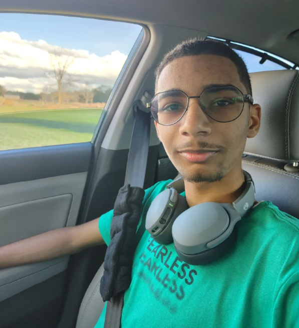 Young man sitting in a car, wearing glasses, headphones around his neck, and a teal T-shirt. The window shows a landscape with green grass, trees, and a partly cloudy sky.