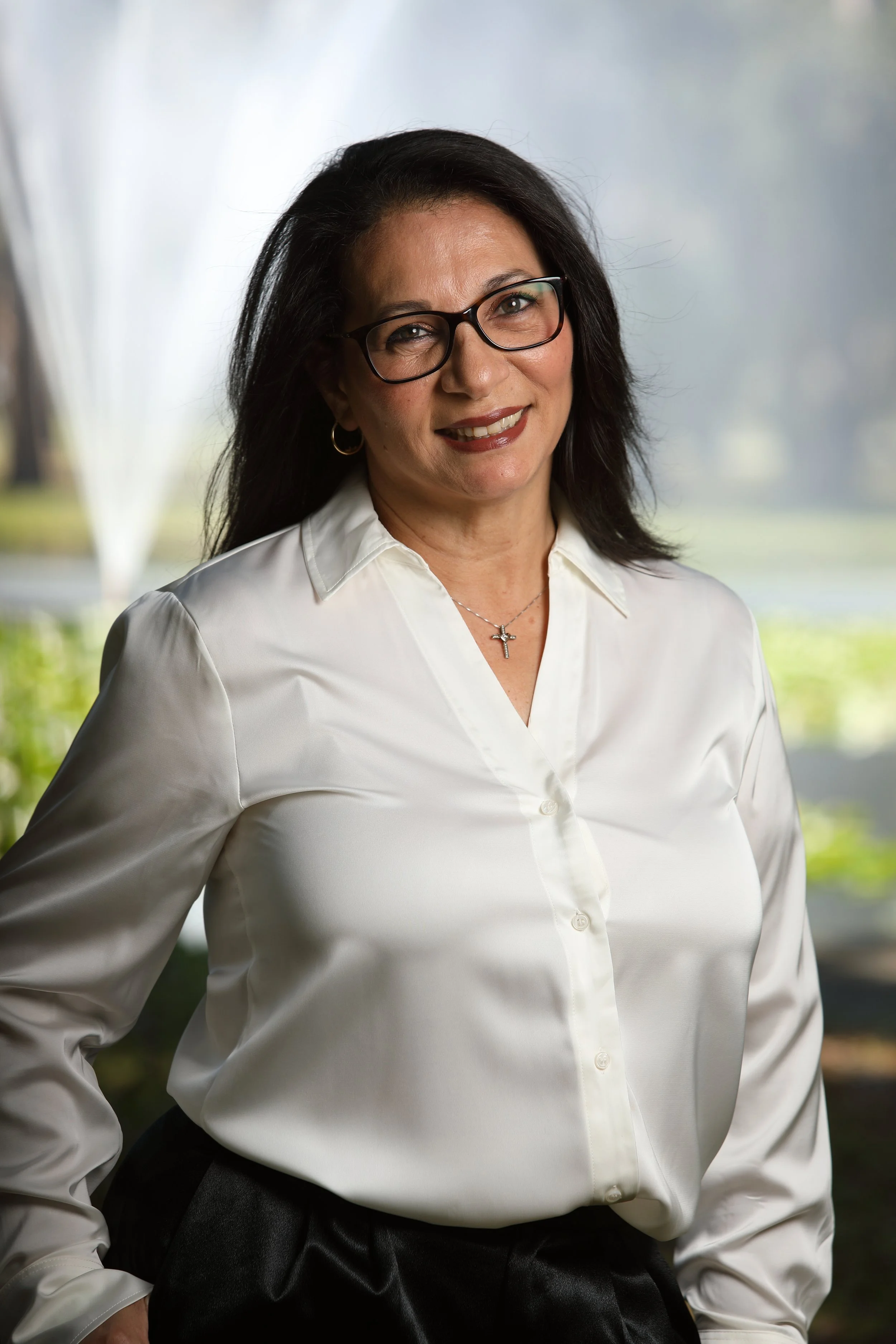 A woman with dark hair and glasses smiling, wearing a white blouse and black pants, standing outdoors with a fountain and green foliage in the background.