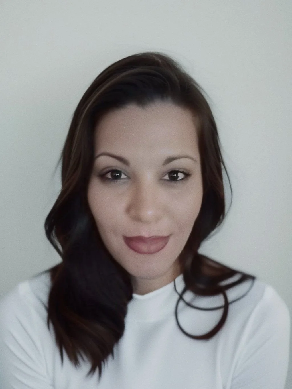 A woman with shoulder-length dark brown hair with lighter highlights, wearing a white top, smiling softly against a plain off-white background.