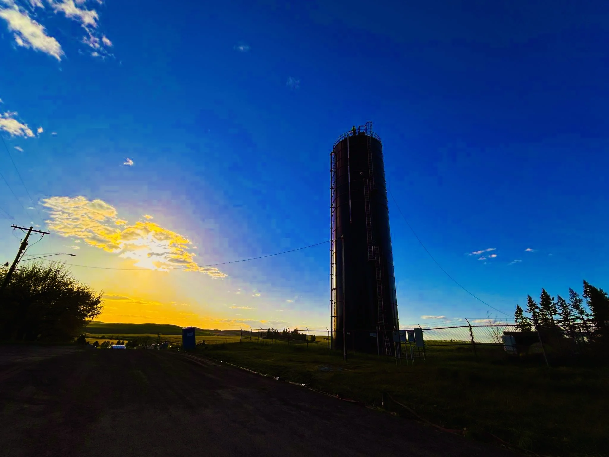 A tall water tower climbed by one of Emerald Creek's operators with a ladder and railings stands against a vibrant sunset sky with clouds. Power lines and a fence are visible in the foreground, along with trees and open land.