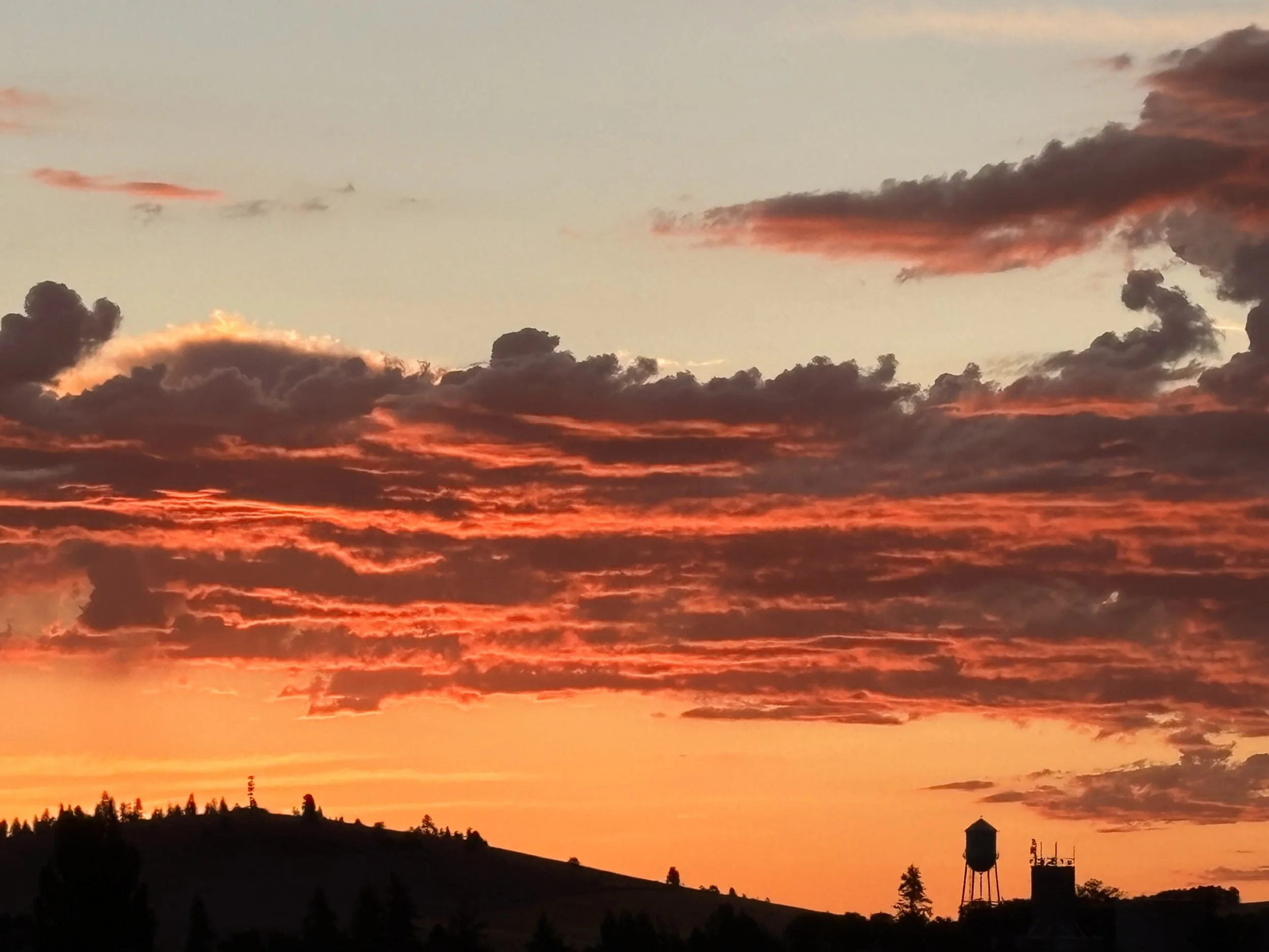 Sunset sky with orange and pink clouds over a silhouette of hills and trees. Water Tower on the horizon take by Emerald Creek.