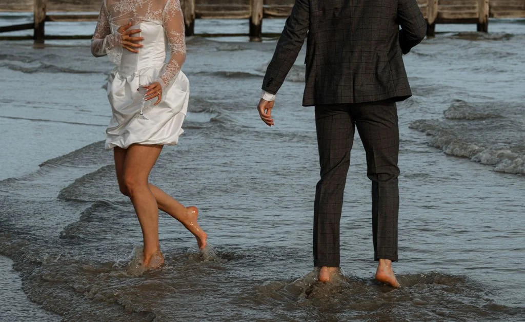 A woman in a white dress and a man in a suit walking barefoot in the water at the beach.