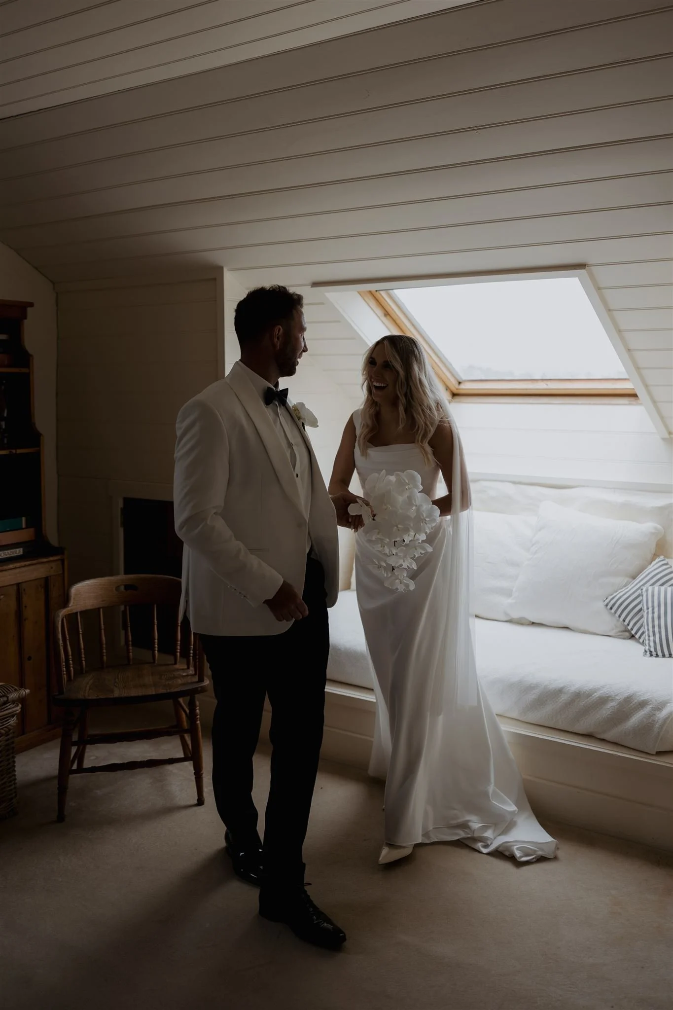 A bride and groom in wedding attire smiling and holding hands in a cozy attic room with a slanted ceiling and a skylight window.
