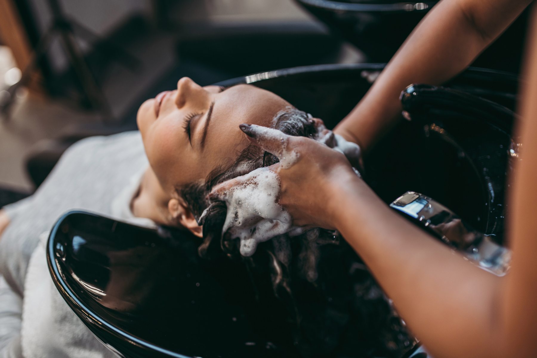 Woman receiving a hair wash at a salon, head reclined over a black sink with shampoo and soap lather on her hair and hands.