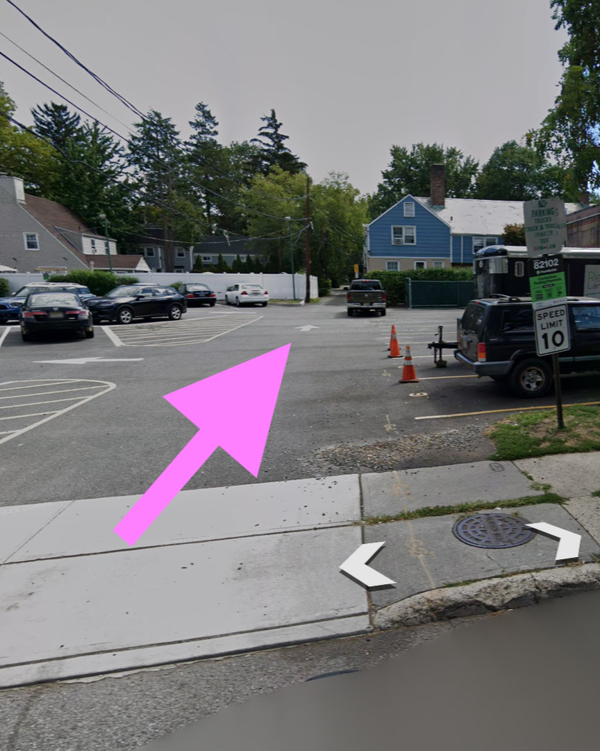 A parking lot with several cars parked and moving, some orange traffic cones, a black vehicle with a trailer with equipment, a sign with speed limit 10, and residential houses in the background.