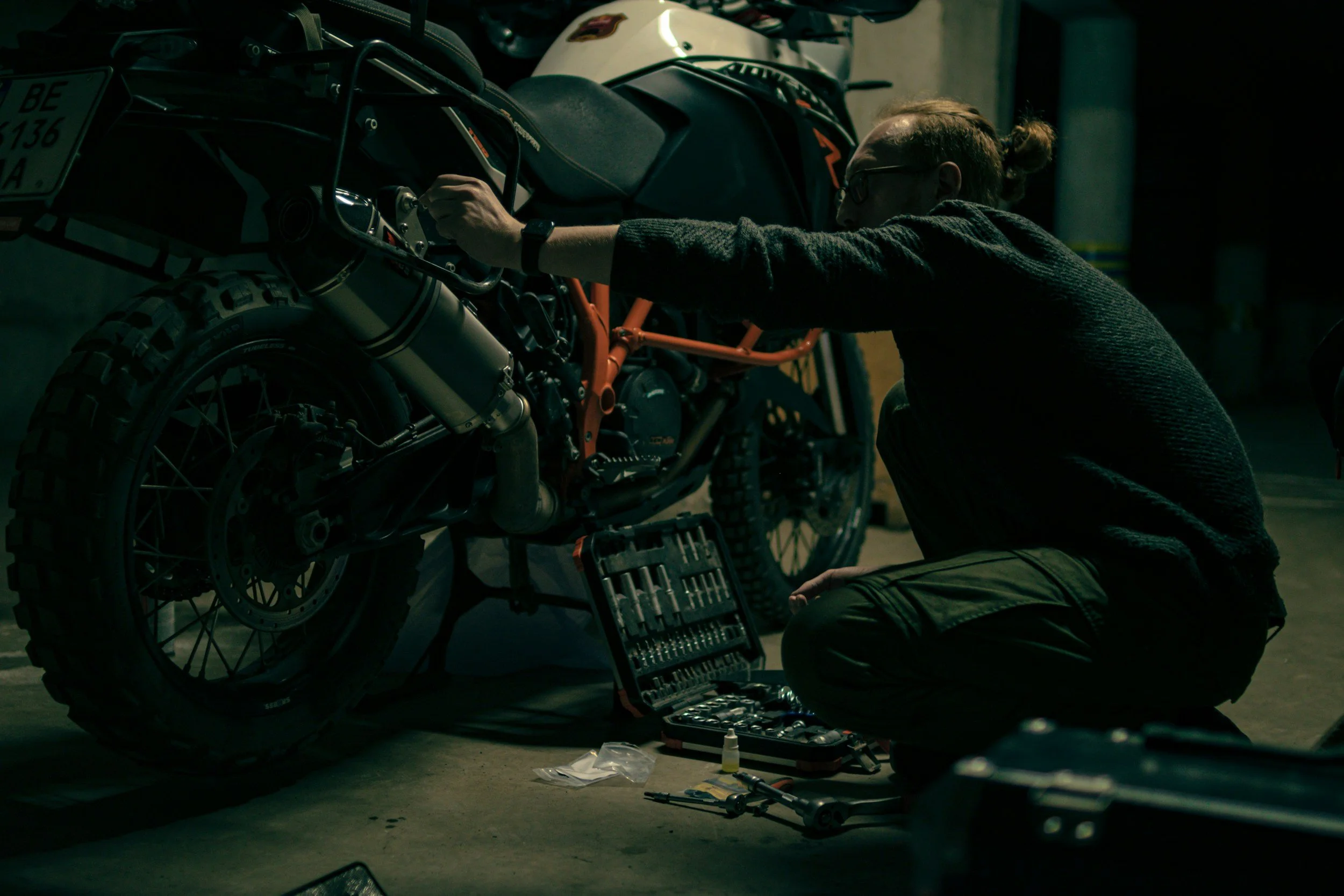 A person working on a motorcycle in a dimly lit garage, kneeling with tools and a toolbox nearby.