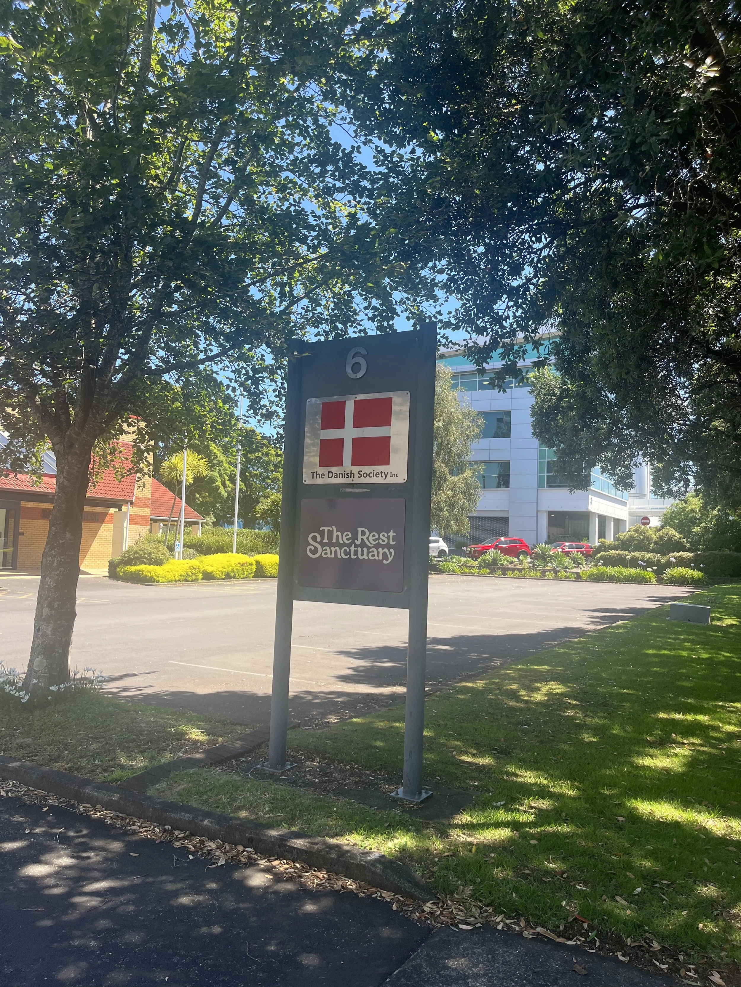 Outdoor sign with The Rest Sanctuary and Danish Society logos, with a carpark behind. A shady tree and green grass are beside the sign.