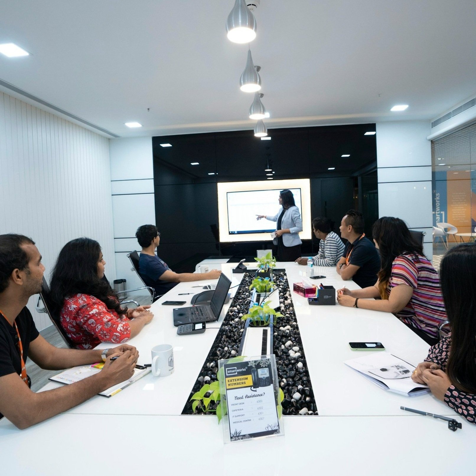 Office meeting with a presenter pointing at a screen in a conference room, featuring a long table and several people seated attentively.