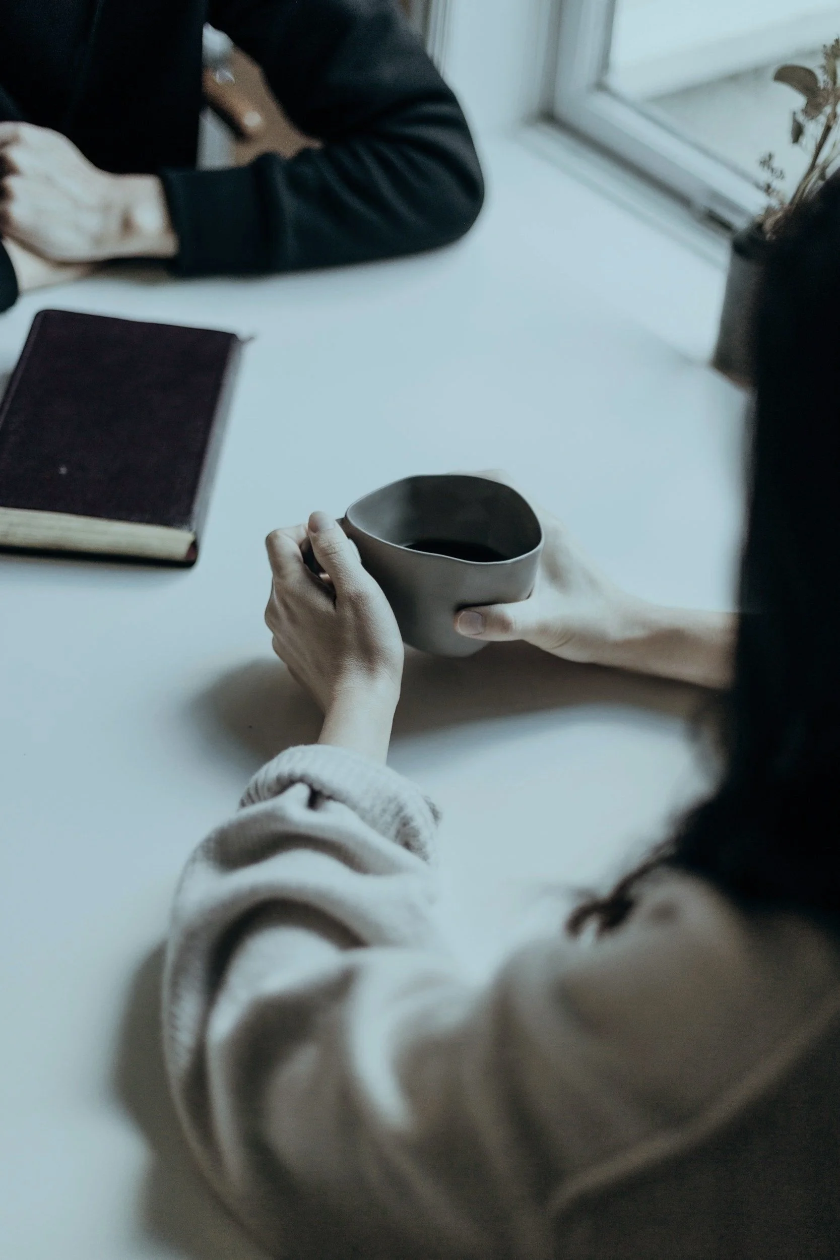Person holding a ceramic cup filled with a dark beverage, sitting at a white table near a window, with a closed book and another person's arm visible on the table.