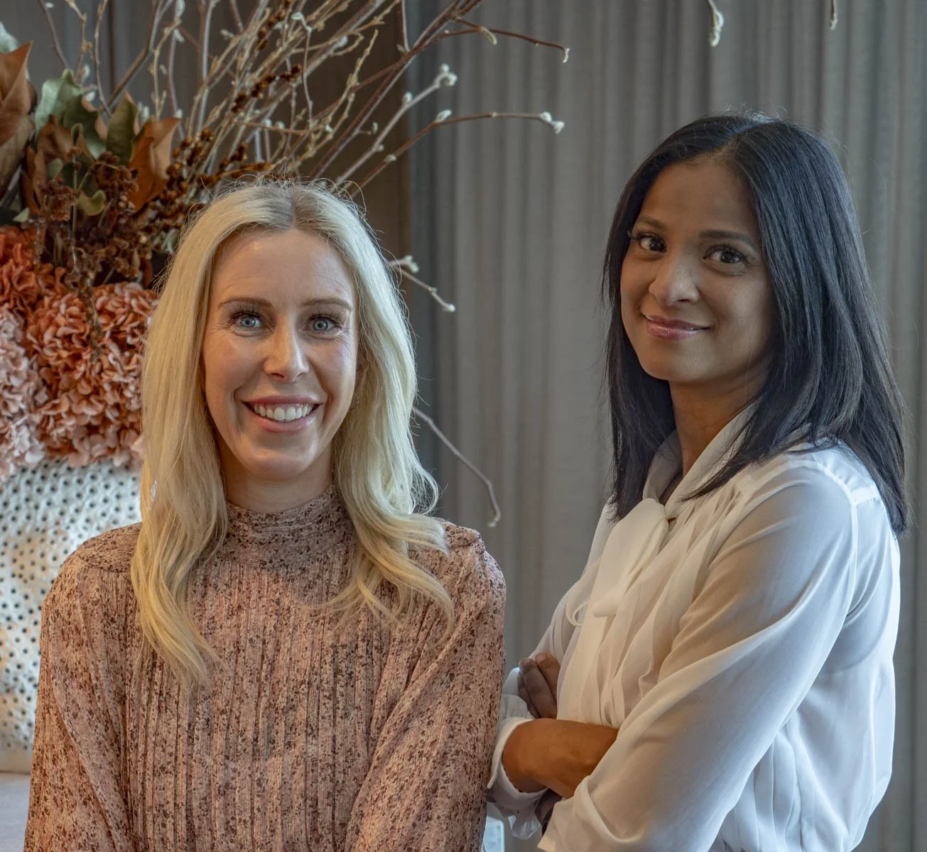 Two women standing indoors near a large floral arrangement, smiling at the camera.