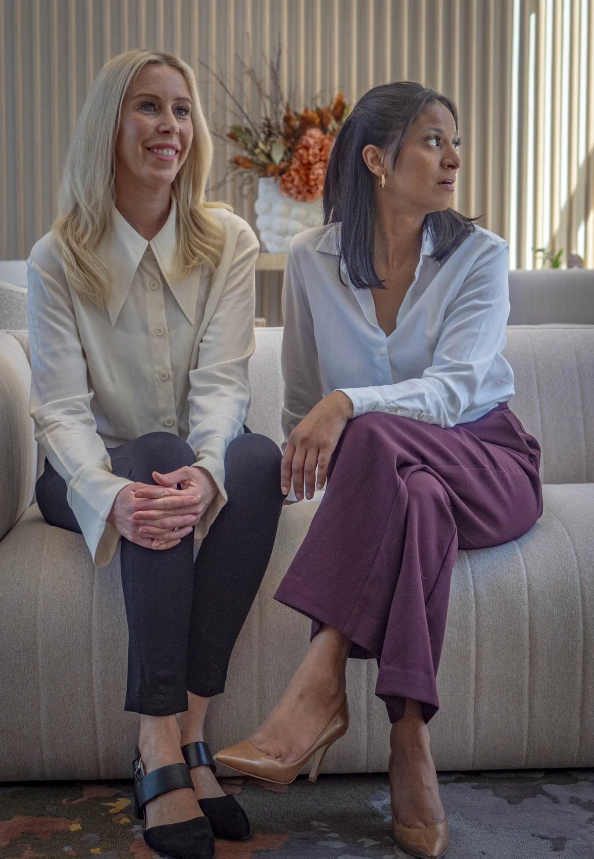 Two women sitting on a beige couch in a modern room with wood paneling and a flower arrangement in the background. One woman has blonde hair, is smiling, and wears a cream blouse and black pants. The other woman has dark hair, wears a white blouse and purple pants, and appears to be looking away.