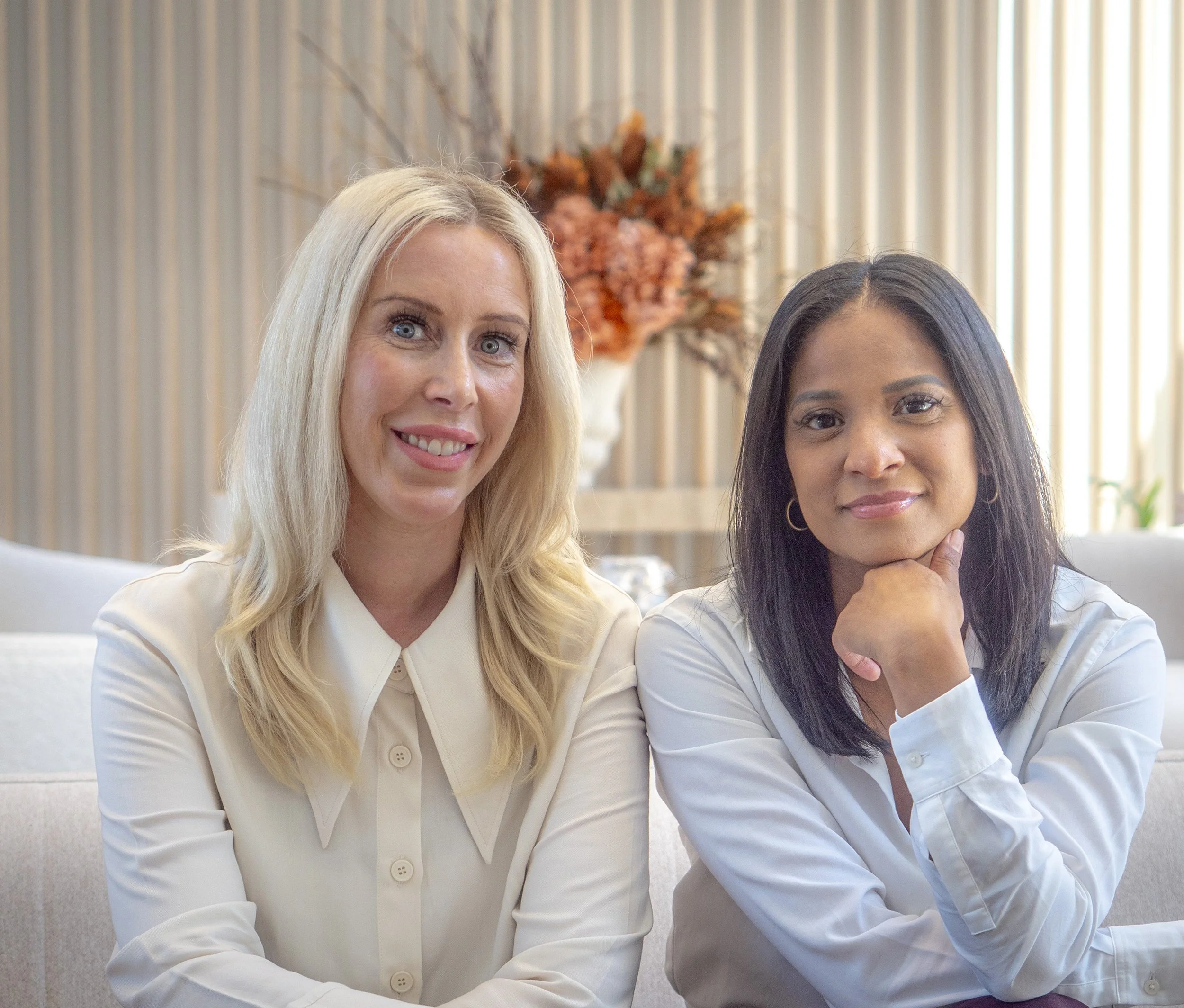 Two women sitting on a beige sofa smiling, with a floral arrangement in the background.