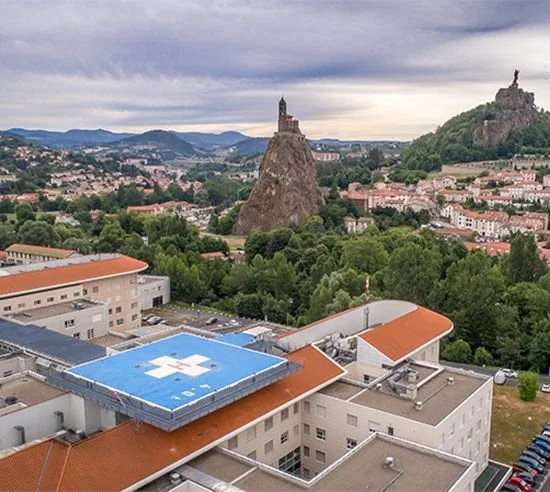 A cityscape featuring two prominent rock formations with historical statues on top, surrounded by green trees and modern buildings, under a cloudy sky.