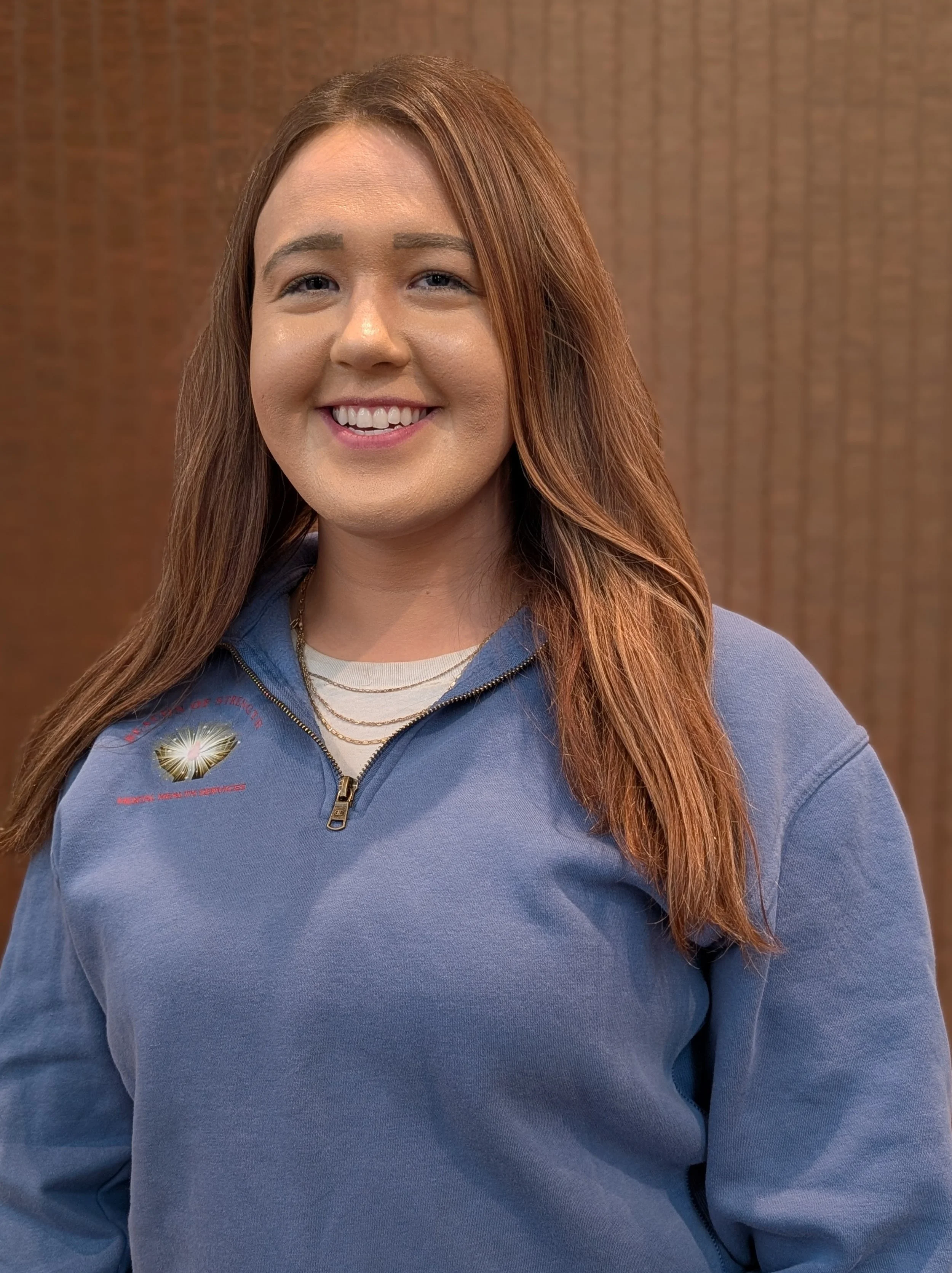 A smiling woman with long, wavy light brown hair, light skin, and blue eyes, wearing a black vest over a beige textured top, with a gold necklace, in an indoor setting.
