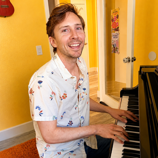 A smiling young man with short brown hair and wearing a white shirt with flowers. He is sitting in front of a piano.