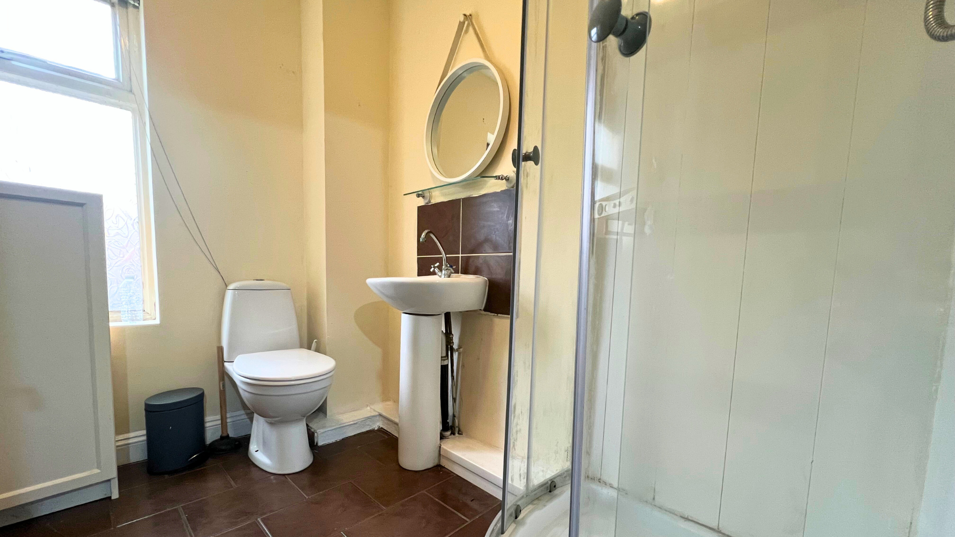 A small bathroom with a toilet, a pedestal sink, a mirror, and a shower with a glass door. The room has beige walls, a window, and brown tiled flooring.